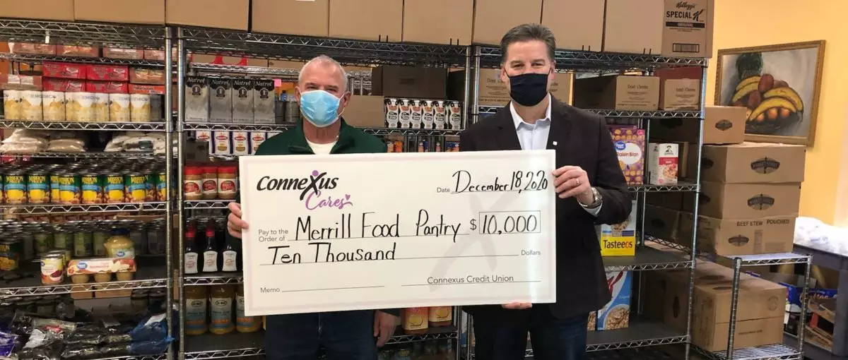 Two people standing indoors in a food pantry. They are each holding one end of a presentation check for $10,000 made out to Merrill Food Pantry.