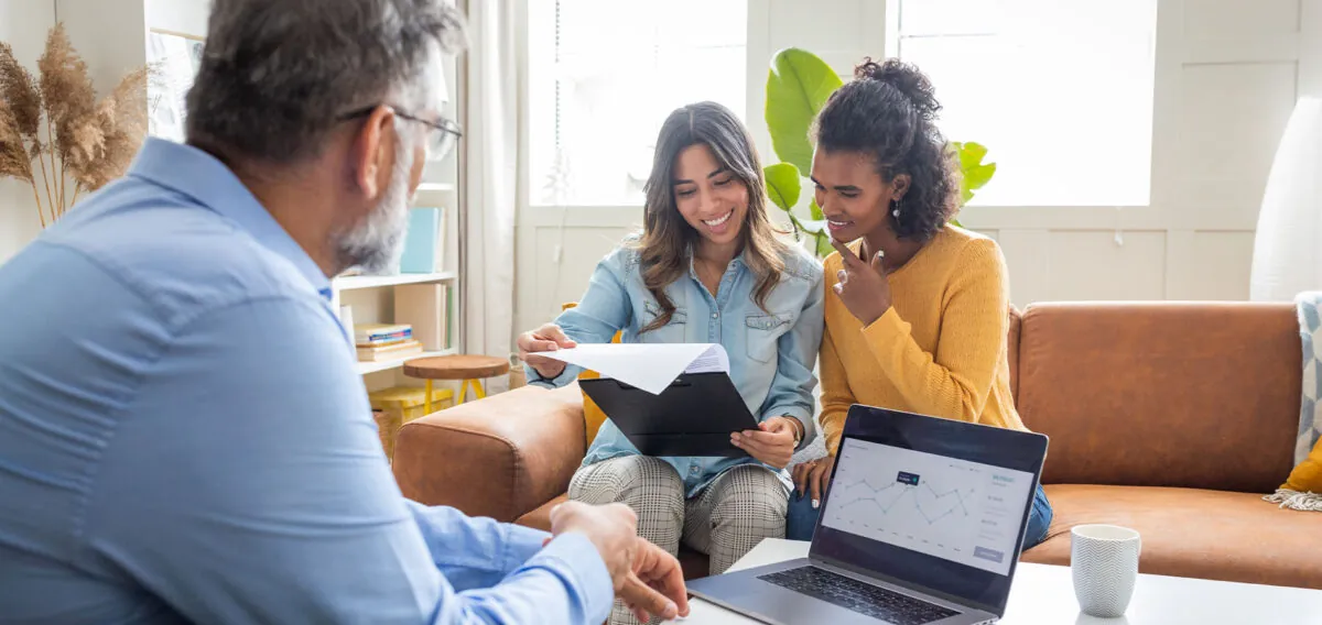 Two women on a couch reviewing paperwork. A man sits across from them working on a laptop.